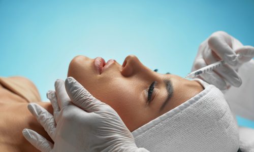 Close up of botox injection in female forehead. Side view of cosmetologist using syringe with special liquid, holding chin while patient with towel on head lying. Concept of cosmetology, beauty.